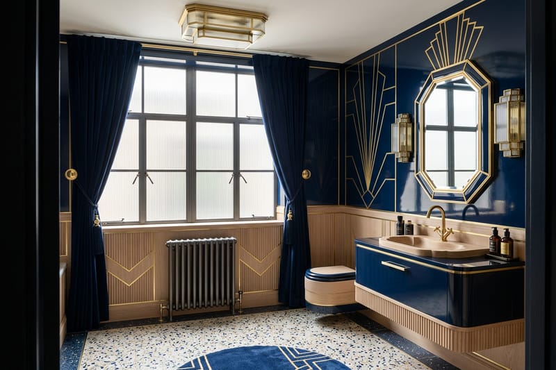 The same room renovated as an Art Deco bathroom in navy and brass, with fluted panelling and terrazzo flooring, keeping the cast-iron radiator