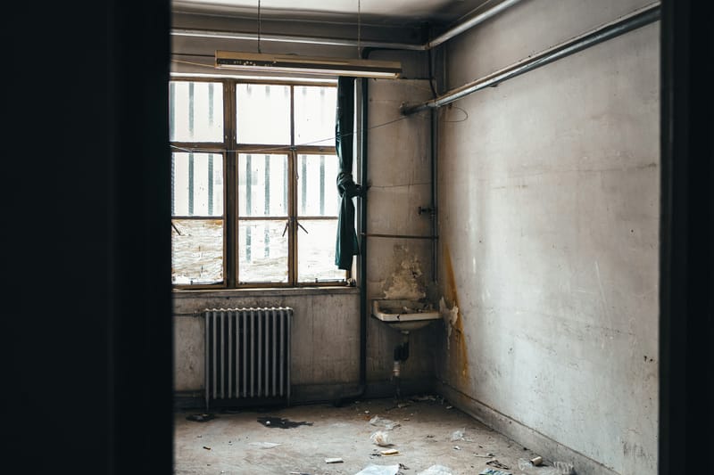A narrow derelict room with exposed pipework, a cracked wall-hung sink and a cast-iron radiator under the window